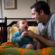 An image of two children happily playing with toys in a daycare center, while a group of dads sit and chat in the background.